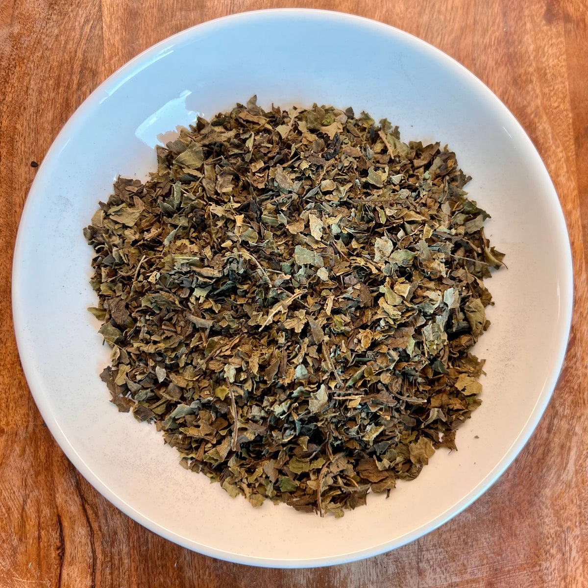 Dried Black Walnut tea leaves piled in a white ceramic bowl, set on a round wooden serving board.