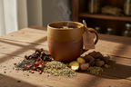 Dandelion & Thistle herbal tea in a rustic ceramic mug surrounded by dandelion root, milk thistle seeds, fennel seeds, ginger root, slippery elm bark and burdock root on a wooden surface