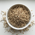 Dried nettle root cut and sifted — thin fibrous stringy pieces in a white ceramic bowl with some scattered on white surface